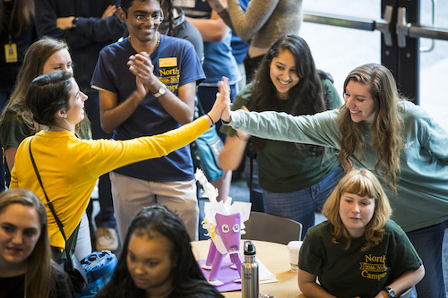 Students high five across a table in a room full of people